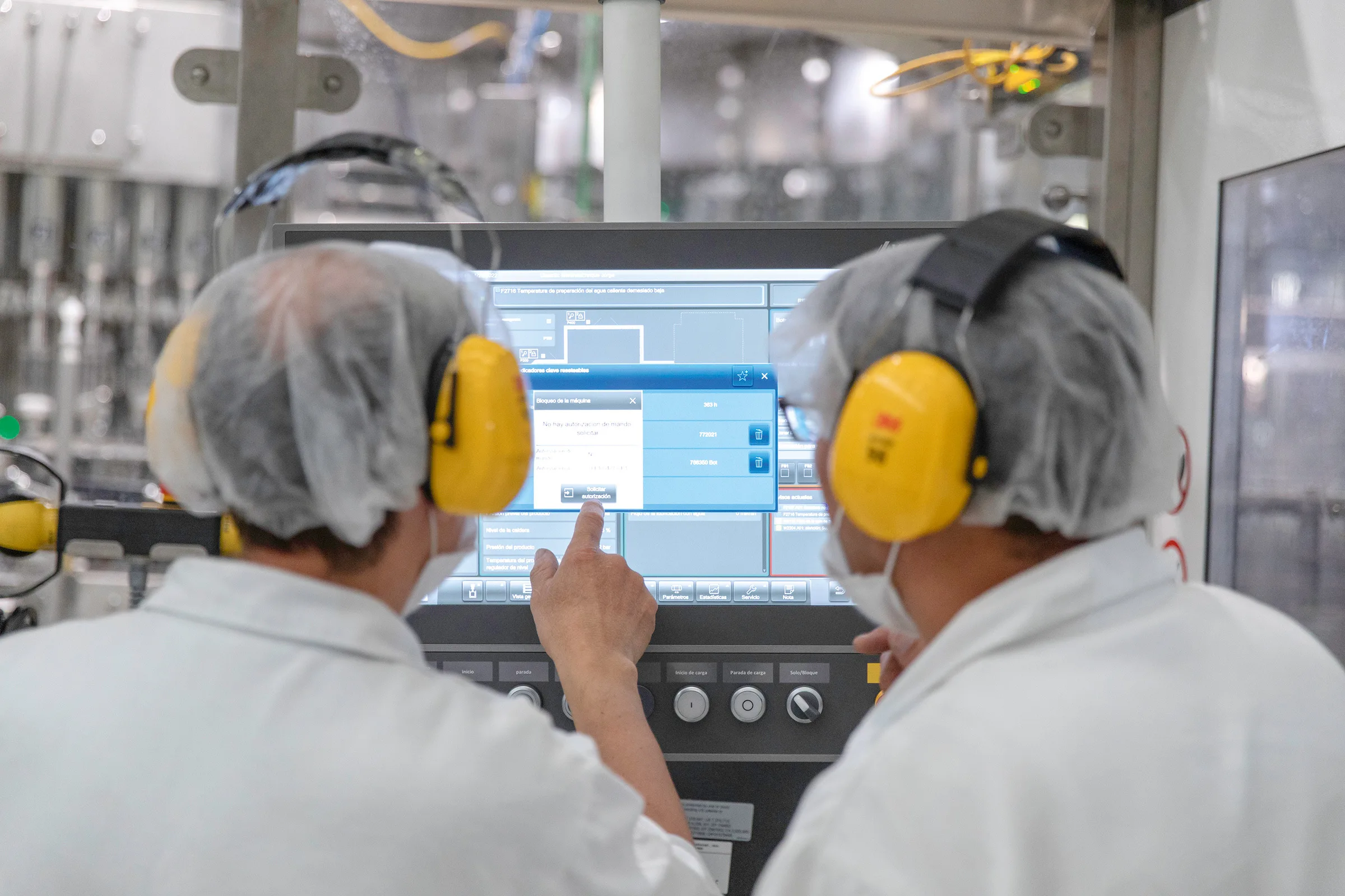 Zwei Personen mit gelbem Gehörschutz und in weißen Kitteln, die auf einen Bildschirm an einer Maschine schauen
